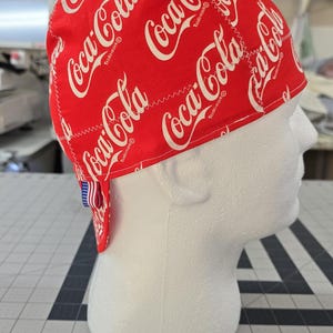 May include: A red scrub cap featuring a repeating white Coca-Cola logo pattern. The fabric head covering has a rounded crown and a small fabric tag with an American flag design attached to the back edge, displaying the brand name clearly.
