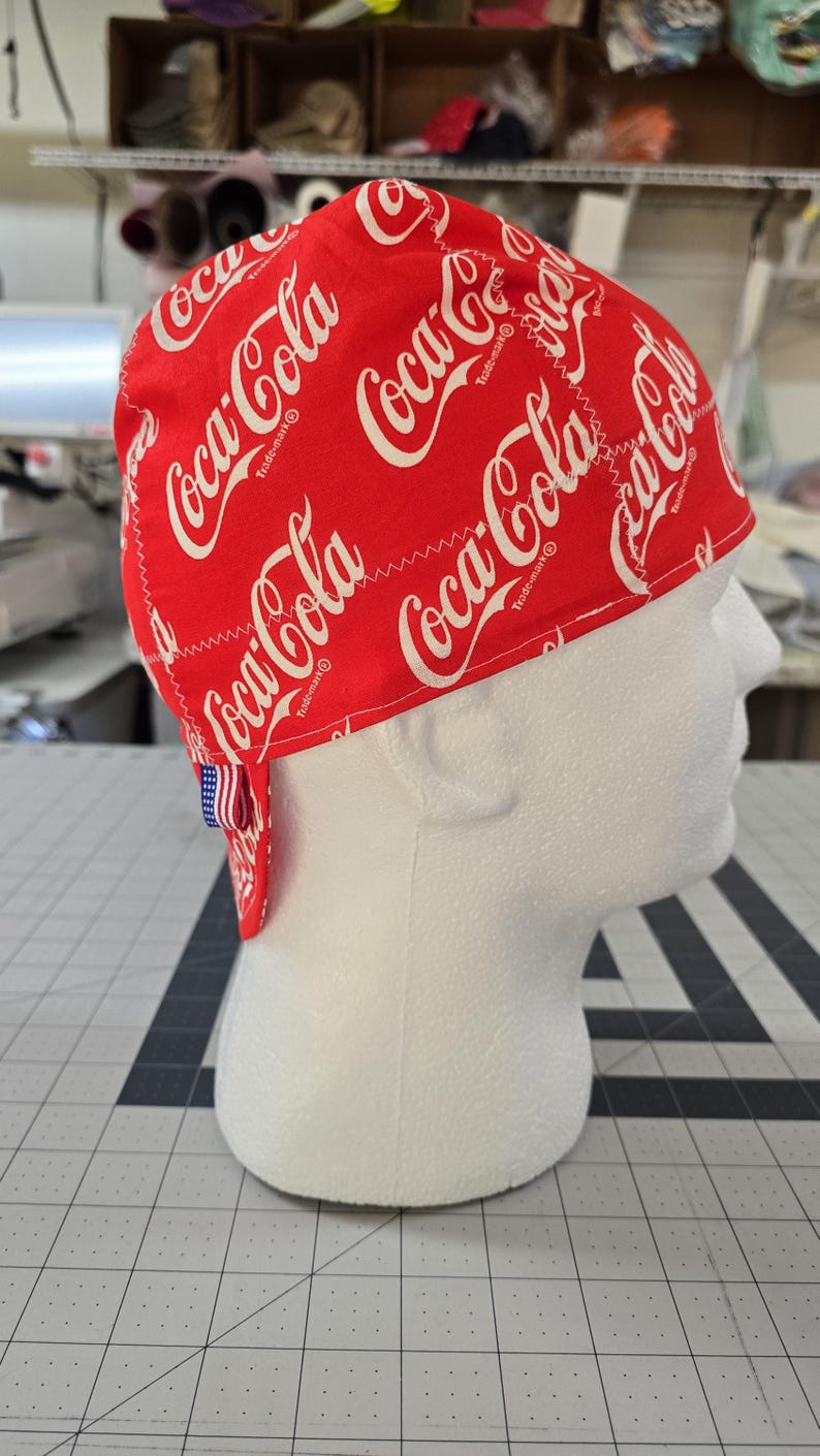 May include: A red scrub cap featuring a repeating white Coca-Cola logo pattern. The fabric head covering has a rounded crown and a small fabric tag with an American flag design attached to the back edge, displaying the brand name clearly.