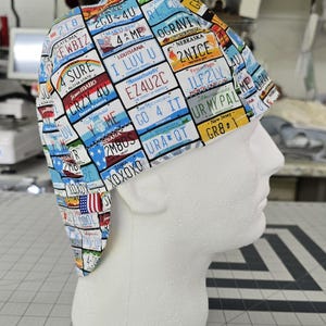 May include: A scrub cap featuring a colorful all-over print of various United States license plates. The fabric displays multiple state names and alphanumeric combinations such as 2GD 4U, I LUV U, EZ4U2C, 2NICE, 1LF2LV, UR MY PAL, GO 4 IT, and XOXOXO.