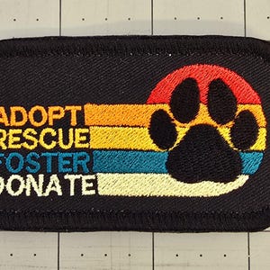 May include: Black embroidered patch with a rainbow stripe design and a paw print. The text "Adopt Rescue Foster Donate" is embroidered in white.