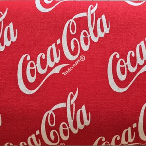 May include: A red fabric featuring a repeating white Coca-Cola logo pattern. The iconic script text is printed in a diagonal orientation across the surface, with each logo including the small trademark symbol next to the word Coca-Cola.