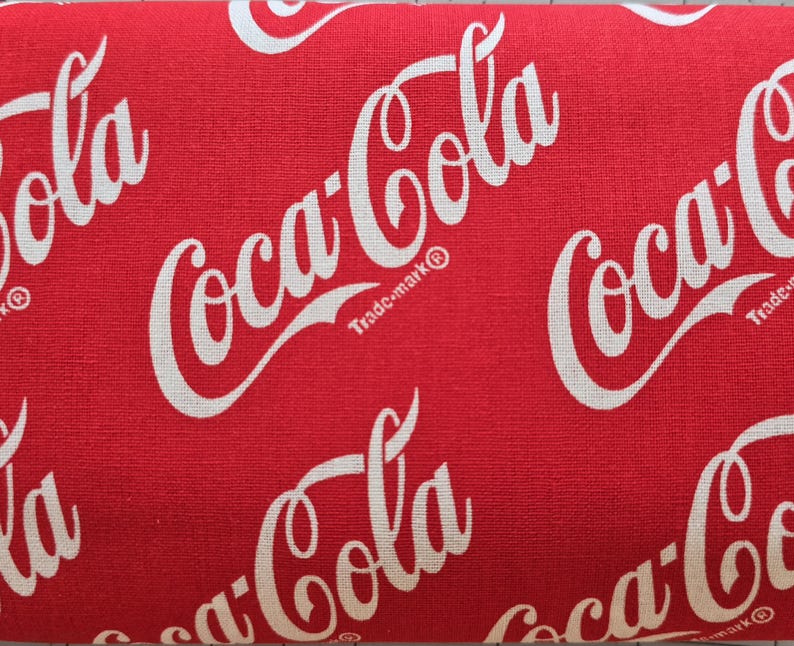 May include: A red fabric featuring a repeating white Coca-Cola logo pattern. The iconic script text is printed in a diagonal orientation across the surface, with each logo including the small trademark symbol next to the word Coca-Cola.