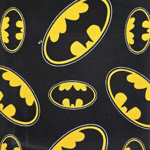 May include: A black fabric featuring a repeating pattern of yellow Batman logos outlined in white. Each oval-shaped emblem displays the iconic bat silhouette in black against a yellow background, with a small TM symbol visible near one of the logos.