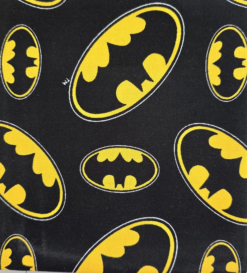 May include: A black fabric featuring a repeating pattern of yellow Batman logos outlined in white. Each oval-shaped emblem displays the iconic bat silhouette in black against a yellow background, with a small TM symbol visible near one of the logos.