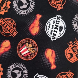 May include: A black fabric featuring a repeating pattern of orange chicken drumsticks and circular badges. The badges contain text including "Keep Calm and Eat Wings," "I'm Just Here for the Wings," and "Had Me at Wings," along with chicken illustrations.