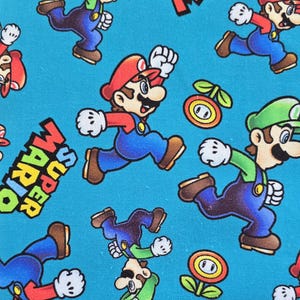 May include: A bright blue fabric featuring a repeating pattern of Mario and Luigi characters in their signature red and green outfits, along with orange fire flower icons and the text Super Mario printed in bold black and yellow letters.