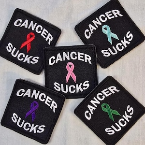 May include: Five black square patches with the words "CANCER SUCKS" embroidered in white. Each patch features a different colored ribbon: red, light blue, pink, purple, and green. The patches are arranged on a light-colored surface.