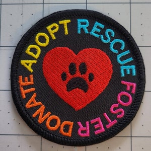 May include: Round black embroidered patch with the words "ADOPT RESCUE FOSTER DONATE" in rainbow colors. A red heart with a black paw print is in the center. The patch is for animal rescue.