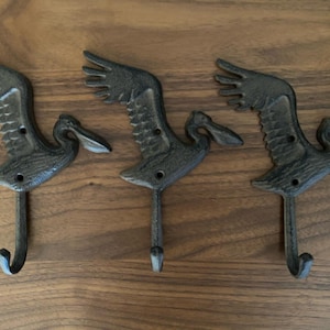 May include: Three black cast iron pelican hooks. Each hook is shaped like a pelican with its wings spread out. The hooks are mounted on a wooden surface.