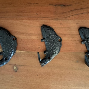 以下が含まれることがあります： 魚の形をした黒い鋳鉄製の壁掛けフック3つ。各フックには詳細な鱗模様、はっきりとした目、フックとして機能する湾曲した尾があります。魚は頭を上、尾を下にして垂直に向けられています。