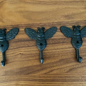 May include: Three cast iron bee-shaped wall hooks. Each hook is black with detailed wings and a downward-facing hook. The hooks are arranged horizontally against a wooden background.