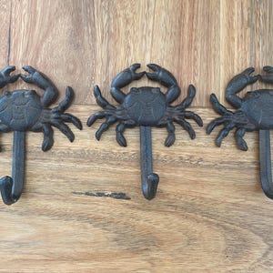 May include: Three cast iron crab-shaped wall hooks. Each hook is a dark, matte grey colour and features a detailed crab design with outstretched claws and a curved hook below. The hooks are mounted on a wooden surface.