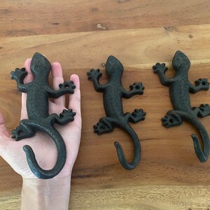 May include: Three black cast iron lizard hooks. The lizards are in a walking pose with their tails curled into a hook shape. The hooks are mounted on a wooden surface.