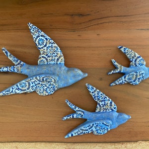 Puede incluir: Tres decoraciones de pared de golondrina de cerámica azul con patrones florales blancos y dorados. Las golondrinas están en vuelo y son de diferentes tamaños.