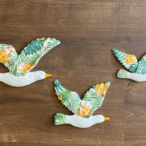 以下が含まれることがあります： 熱帯の葉と花の模様が施された白いカモメの壁飾り3つ。カモメはサイズが異なり、異なる方向に飛んでいます。