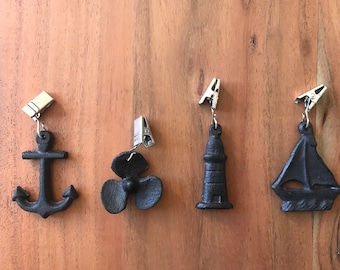 May include: Four nautical-themed decorative clips. The clips feature an anchor, propeller, lighthouse, and sailboat, all in a dark, matte finish. Each clip is attached to a silver-colored metal clip, set against a wooden background.