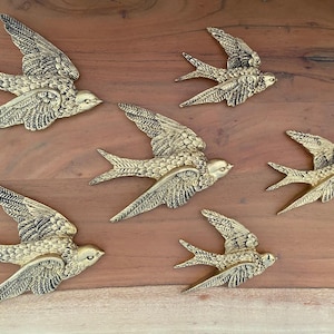 May include: Six gold-colored swallow bird wall decorations with detailed wing and feather designs. The birds are arranged on a wooden surface, creating a sense of flight and movement. The birds are made of metal.