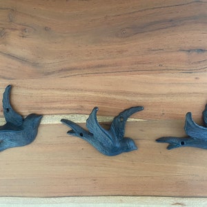 May include: Three black cast iron bird hooks mounted on a wooden surface. The birds are in flight position with their wings spread out.