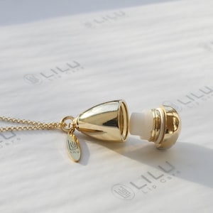 May include: A gold necklace with a gold pendant shaped like a teardrop. The pendant is open and reveals a white lip balm inside. The pendant has the word "LILU" engraved on it.