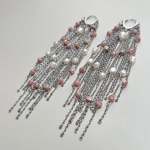 Puede incluir: Un par de pendientes largos colgantes con múltiples cadenas de tono plateado suspendidas de un aro circular. Las cadenas están adornadas con pequeñas cuentas facetadas de color rosa empolvado y perlas de agua dulce blancas irregulares.