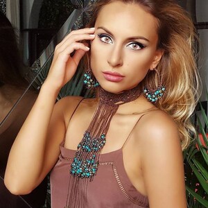 Puede incluir: Un collar de macramé marrón, gargantilla y pendientes de aro a juego con cuentas turquesas. Las joyas se llevan con un top marrón de tirantes finos. Los pendientes y el collar tienen un estilo bohemio.