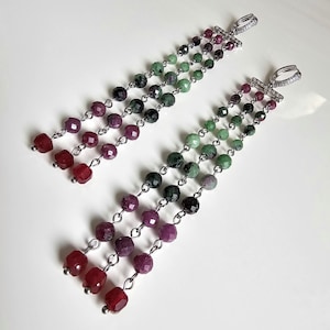 Puede incluir: Un par de pendientes colgantes con múltiples hilos de cuentas facetadas en tonos rojos, morados, verdes y negros. Los pendientes tienen detalles metálicos plateados y un pequeño aro en la parte superior. Se muestran sobre una superficie blanca.