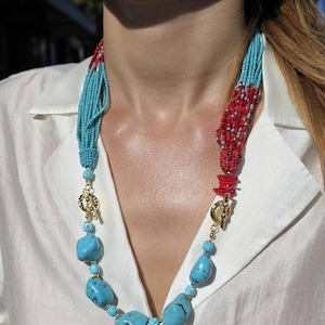 Puede incluir: Un collar de cuentas turquesas y rojas con detalles dorados. El collar presenta piedras turquesas grandes e irregulares y cuentas turquesas más pequeñas, con secciones de cuentas rojas. El collar se abrocha con un cierre dorado.