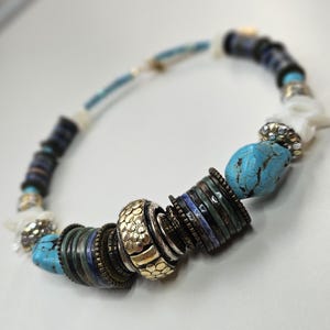 Puede incluir: Un collar de cuentas con piedras turquesas, discos de color azul oscuro y verde, espaciadores metálicos dorados con textura y detalles blancos similares a conchas. El collar está ensartado con una variedad de cuentas cilíndricas.