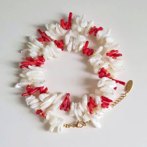 Puede incluir: Un collar con piezas de concha blanca intercaladas con vibrantes acentos de coral rojo. El collar tiene una cadena y un cierre de color dorado, creando un contraste llamativo. El diseño es de estilo natural y orgánico.