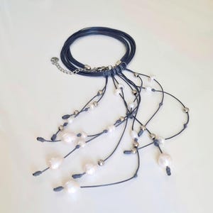 Puede incluir: Un collar azul marino con múltiples hebras de cordón azul oscuro. El collar está adornado con perlas blancas y pequeñas cuentas plateadas, creando un diseño elegante y en capas. El collar tiene un cierre plateado.