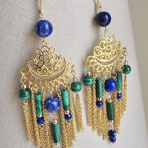 Puede incluir: Un par de pendientes colgantes dorados. Cada pendiente presenta un diseño de filigrana, cuentas de piedras preciosas azules y verdes y cadenas doradas colgantes. Cierre de gancho.
