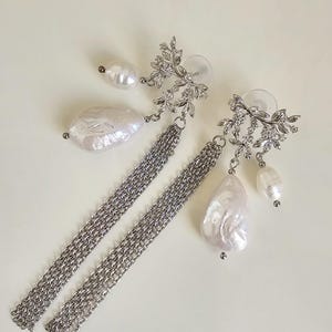 Puede incluir: Elegantes pendientes plateados con diseño floral, cadenas colgantes y perlas blancas brillantes. Los pendientes tienen cierre de poste. Las perlas varían en tamaño y forma, añadiendo un toque único.