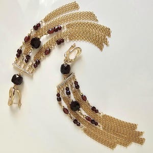 Puede incluir: Pendientes colgantes dorados con detalles de piedras preciosas rojo oscuro. Los pendientes combinan borlas de cadena, pequeñas cuentas y piedras preciosas facetadas más grandes. Cierre de palanca. Aproximadamente 8 cm de largo.