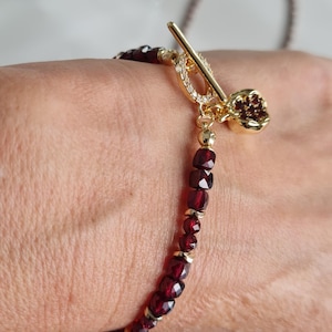 Op de afbeelding: Een gouden armband met een hartvormige hanger en een schuifgesp. De armband is gemaakt van kleine, gefacetteerde rode edelstenen.