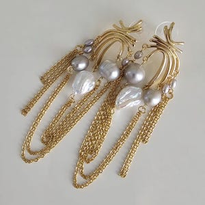 Puede incluir: Pendientes dorados con un diseño único. Cada pendiente presenta una parte superior dorada curvada, perlas grises y blancas y cadenas doradas colgantes. Los pendientes tienen un cierre de poste.