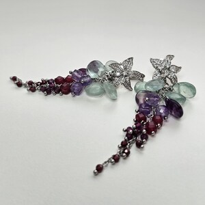 Puede incluir: Un par de pendientes colgantes con postes en forma de flor plateada incrustados con piedras claras. De cada flor cuelgan racimos de cuentas facetadas en tonos rojo oscuro, morado y verde pálido, dispuestos en una forma larga y cónica.