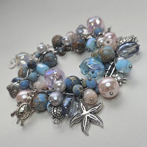Puede incluir: Una pulsera de dijes de múltiples hilos con una variedad de cuentas en tonos azules, rosas y grises. La pulsera incluye dijes de metal plateado con formas de tortugas marinas y una estrella de mar, intercalados con cuentas lisas, facetadas y jaspeadas.