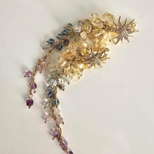 Puede incluir: Dos horquillas doradas con diseños de sol y pedrería transparente. Cada horquilla presenta un grupo de pequeñas cuentas coloridas en tonos amarillos, azules, morados y transparentes, colgando de delicadas cadenas doradas. Accesorios para el cabello.