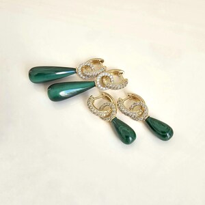 Puede incluir: Pendientes de aro dorados con un diseño brillante y colgantes en forma de lágrima de color verde oscuro. Los pendientes se muestran sobre una superficie blanca, resaltando su elegante diseño y contraste de color.