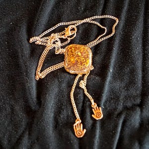 May include: A gold-toned necklace featuring a square pendant with an amber-coloured resin inlay containing swirling patterns. The chain extends into two dangling strands, each ending in a small charm shaped like a hand with a spiral palm design.