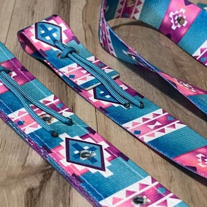 May include: Close-up of several colorful patterned straps. The straps feature a geometric design in shades of turquoise, pink, and purple. Each strap has a turquoise cord and metal eyelets. The straps are likely for a bag or other accessory.