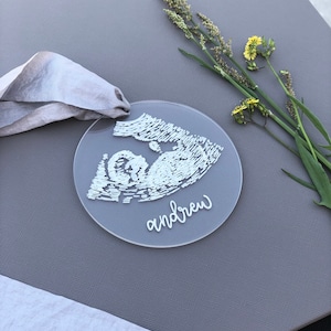 Op de afbeelding: Een ronde, transparante acrylversiering met een witte omtrek van een baby in de baarmoeder. Het woord "andrew" is in witte cursieve letters onder de omtrek geschreven. De versiering ligt op een grijs oppervlak met een grijs lint en een takje gele wilde bloemen.