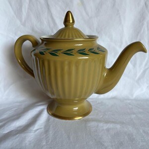 May include: A vintage mustard-yellow teapot with a gold-colored finial and trim. The teapot features a ribbed body and a decorative band of green leaf designs. The handle and spout are curved, and the teapot sits on a small pedestal base.