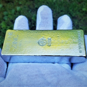 May include: A 10 troy ounces 99.9% fine zinc bar. The bar is silver in color and has the words "99.9% FINE ZINC" and "10 TROY OUNCES" printed on it.