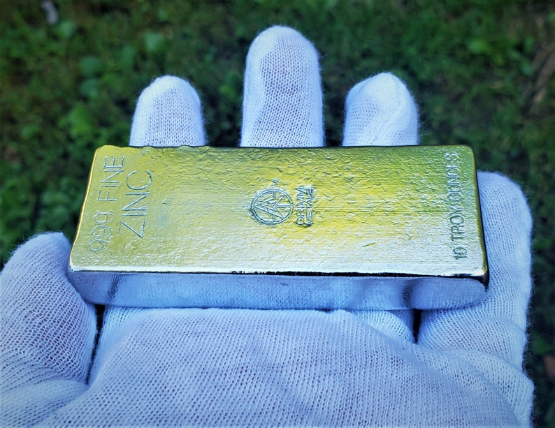 May include: A 10 troy ounces 99.9% fine zinc bar. The bar is silver in color and has the words "99.9% FINE ZINC" and "10 TROY OUNCES" printed on it.
