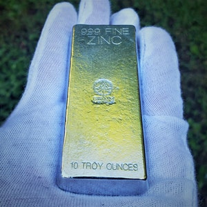 May include: A 10 troy ounce bar of 999 fine zinc. The bar is gold in color and has the words "999 Fine Zinc" and "10 Troy Ounces" printed on it. The bar also has a logo with the words "Grimm Metals" printed on it.