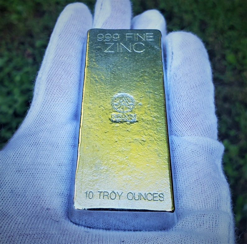 May include: A 10 troy ounce bar of 999 fine zinc. The bar is gold in color and has the words "999 Fine Zinc" and "10 Troy Ounces" printed on it. The bar also has a logo with the words "Grimm Metals" printed on it.