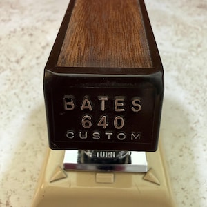May include: A vintage Bates 640 Custom stapler. The stapler features a dark brown wooden top and a cream-colored base. The text "BATES 640 CUSTOM" is embossed on the front. The stapler has a silver metal component with the word "TURN" on it.