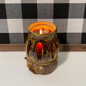 May include: A lit candle in a handcrafted ceramic lantern. The lantern has a rustic design with a brown and green glaze, and a bark-like base. The candle flame glows brightly, casting a warm light through the cut-out design.