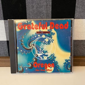May include: A CD case featuring the Grateful Dead album "Oregon: The Day After." The cover art has a blue and yellow fractal design with the band's name in red and the album title in red. A white circle contains a skull and lightning bolt.